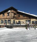 Berghütte aus Holz und Stein im Schnee mit Skiern im Vordergrund unter blauem Himmel.