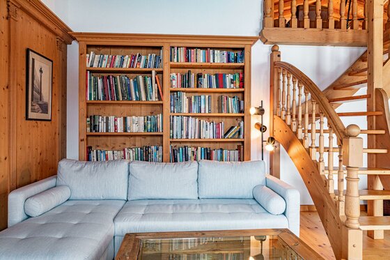 Wohnbereich mit hellem Sofa, Holzregal voller Bücher und einer Holztreppe in einem Chalet.