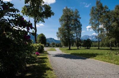 Ganslern Trail Zugangsweg Asphaltierter Weg zwischen Wiesen und Bäumen mit Blick auf Berge im Hintergrund.