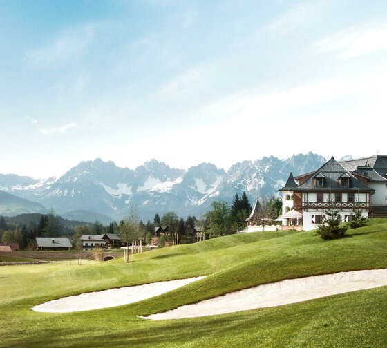 Eine malerische Landschaft mit einem Golfplatz und einem eleganten Gebäude. Im Hintergrund erheben sich majestätische Berge unter einem klaren Himmel.