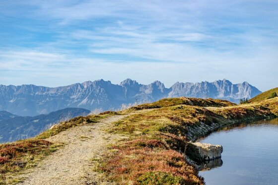 Schotterweg auf einem Graskamm neben einem Bergsee mit Blick auf ein zerklüftetes Gebirgsmassiv unter blauem Himmel.