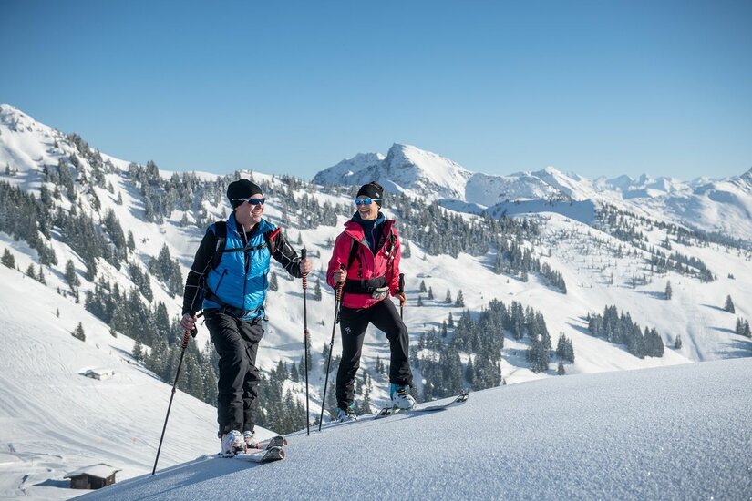 Zwei Skitourengeher auf einem verschneiten Bergrücken vor alpinem Panorama.