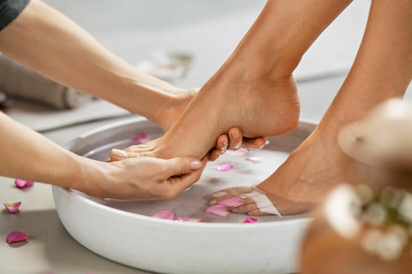 Fußbad und Pediküre Zwei Hände halten einen Fuß über einer weißen Schale mit Wasser und rosa Blütenblättern.