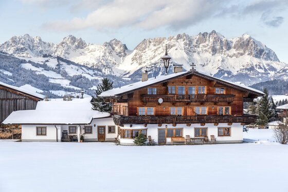 Alpines Holzhaus mit schneebedecktem Dach vor einem verschneiten Gebirgspanorama.