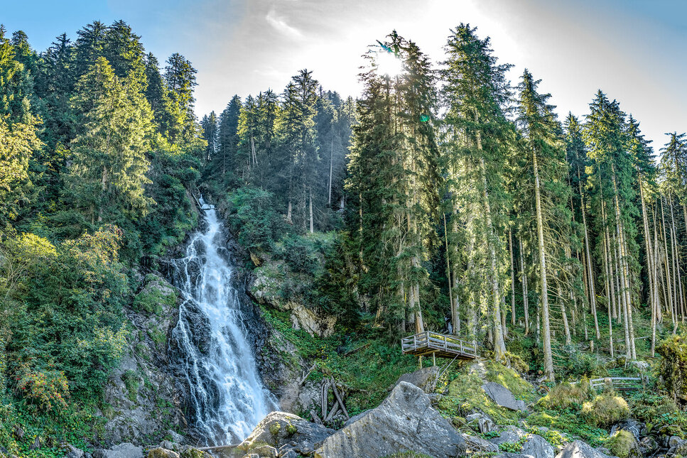 Ein majestätischer Wasserfall stürzt zwischen üppigen, grünen Bäumen in eine felsige Umgebung, während die Sonne strahlt.