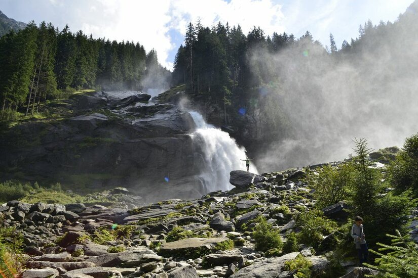 Ein großer Wasserfall stürzt über dunkle Felsen in ein steiniges Becken, umgeben von Nadelwald.