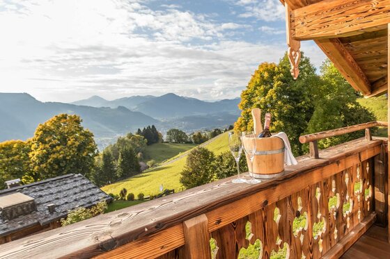 Ausblick von Rosi´s Sonnbergstuben Holzbalkon mit Sektkühler und Gläsern vor einer alpinen Berglandschaft mit grünen Wiesen und Wäldern.