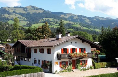 Zweistöckiges weißes Alpenhaus mit roten Fensterläden und Blumenschmuck vor bewaldeten Bergen.