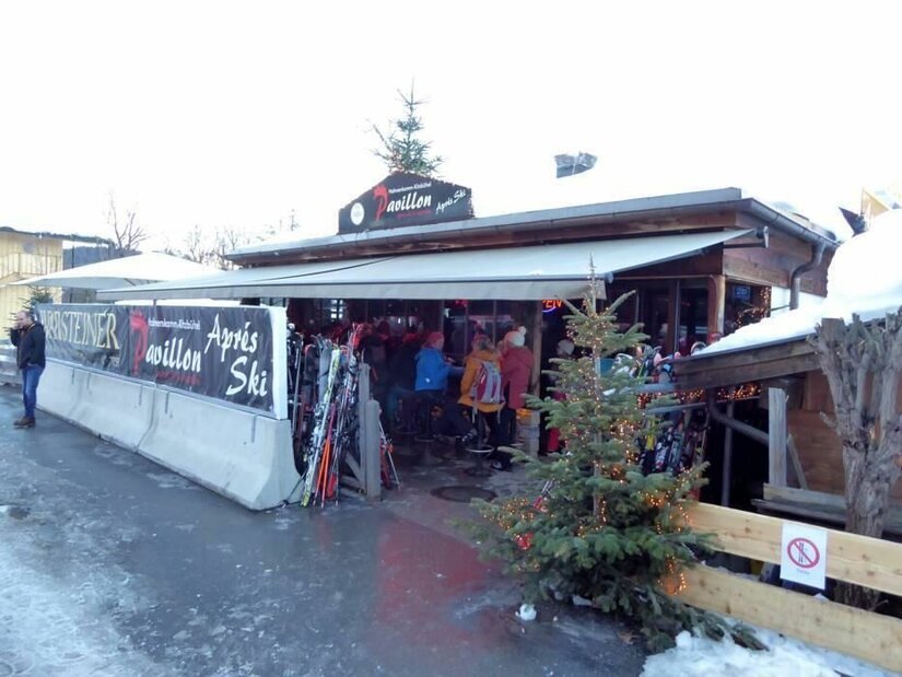 Hahnenkamm Pavillon Außenansicht des Hahnenkamm Pavillons mit Skifahrern und abgestellten Skiern unter einer Markise.
