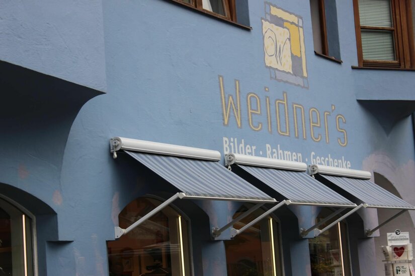 Geschäft Weidner Blaue Hausfassade mit der Aufschrift Weidner und drei gestreiften Markisen über Schaufenstern.