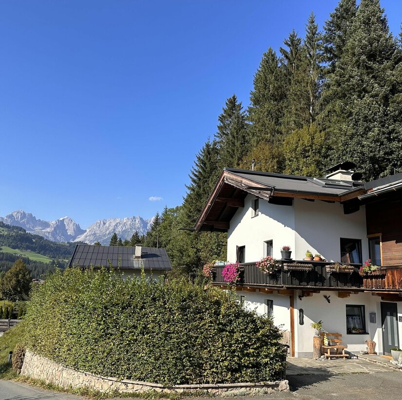 Wohnhaus Tannenheim mit Bergpanorama Alpines Wohnhaus mit Blumenbalkon vor bewaldetem Hang und Gebirgskette unter blauem Himmel.