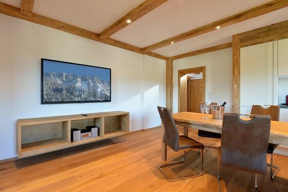Wohnbereich im Pöllhof Moderner Wohnraum mit Holzbalkendecke, Esstisch, Stühlen und einem Fernseher an der Wand.