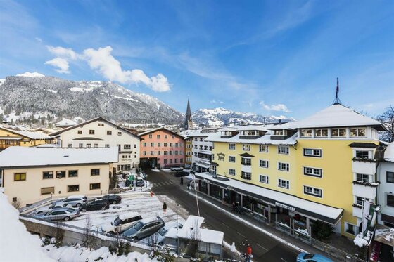 Ortszentrum im Winter Verschneite Ortsansicht mit mehrstöckigen Gebäuden, Parkplatz und Bergen im Hintergrund unter blauem Himmel.