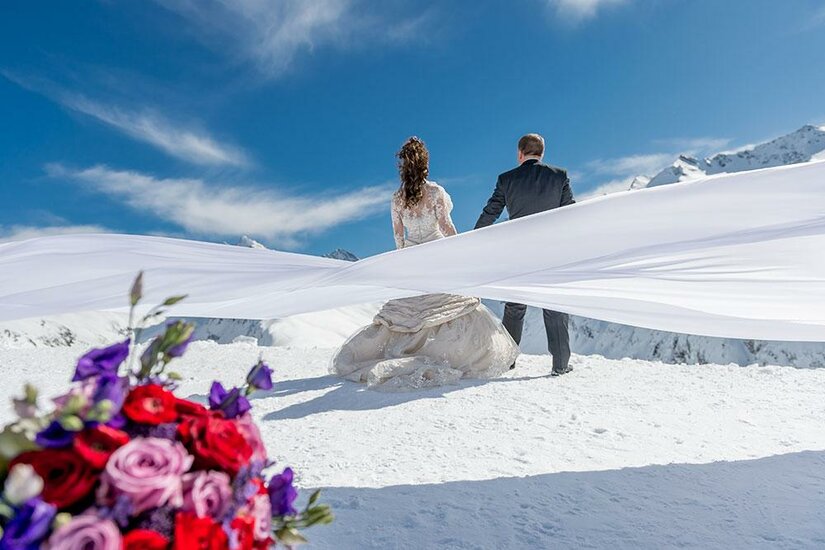 Hochzeit im Schnee Ein Brautpaar steht auf einem schneebedeckten Berg mit atemberaubender Aussicht. Im Vordergrund sind bunte Blumen zu sehen.