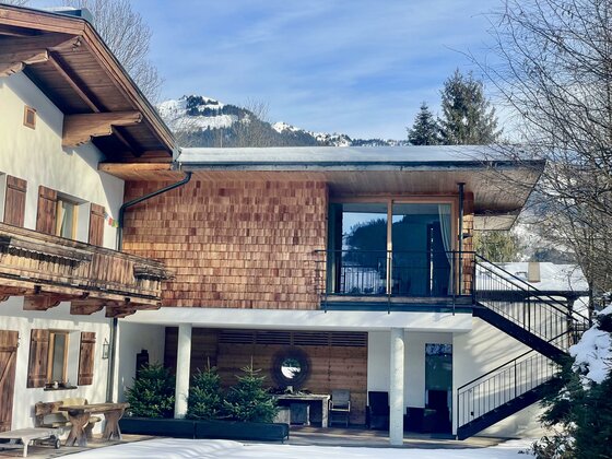 Ein modernes Haus mit Holzverkleidung und großen Fenstern. Im Hintergrund sind schneebedeckte Berge zu sehen.