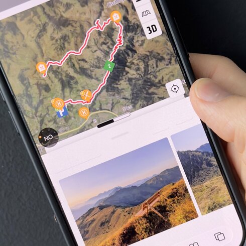 Eine Hand hält ein Smartphone, das eine detaillierte Karte mit Wanderwegen in den Bergen zeigt, umgeben von Natur.