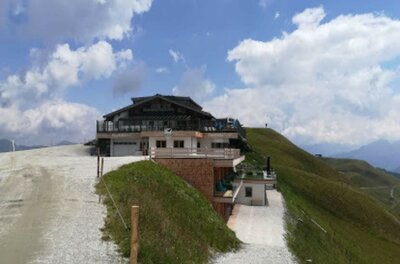 Berghütte mit Holzterrasse an einem Schotterweg vor grasbewachsenen Hängen unter bewölktem Himmel.