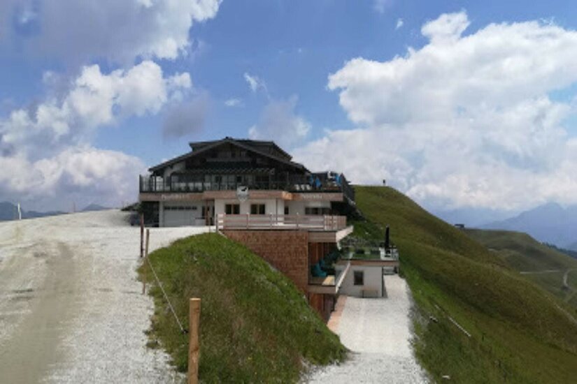 Ehrenbachgraben im Sommer Berghütte mit Holzterrasse an einem Schotterweg vor grasbewachsenen Hängen unter bewölktem Himmel.