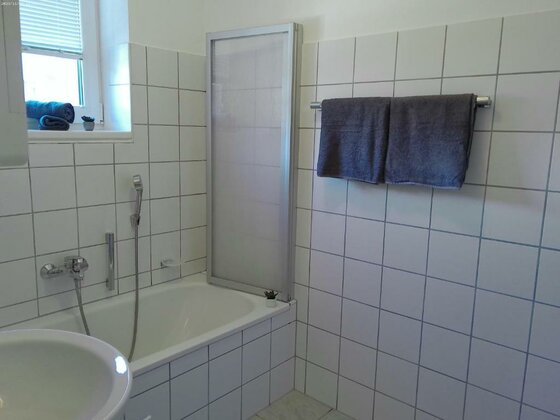 Ein modernes Badezimmer mit einer Badewanne und einer Duschtrennwand. Es ist hell und sauber, mit weißen Fliesen und zwei Handtüchern.