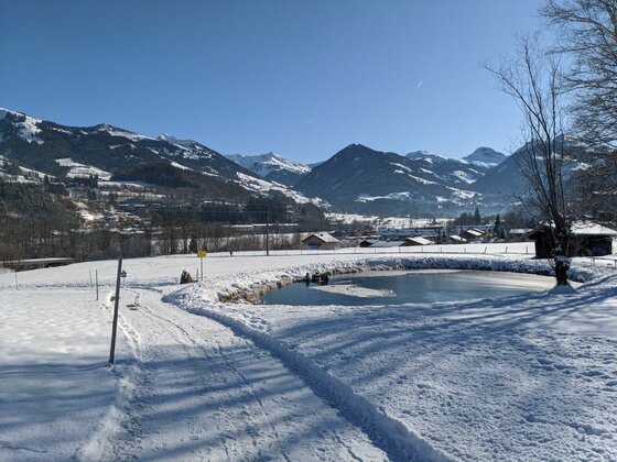 Winterlandschaft bei Malerndörfl Verschneite Landschaft mit einem kleinen Teich, einem Winterwanderweg und schneebedeckten Bergen im Hintergrund.