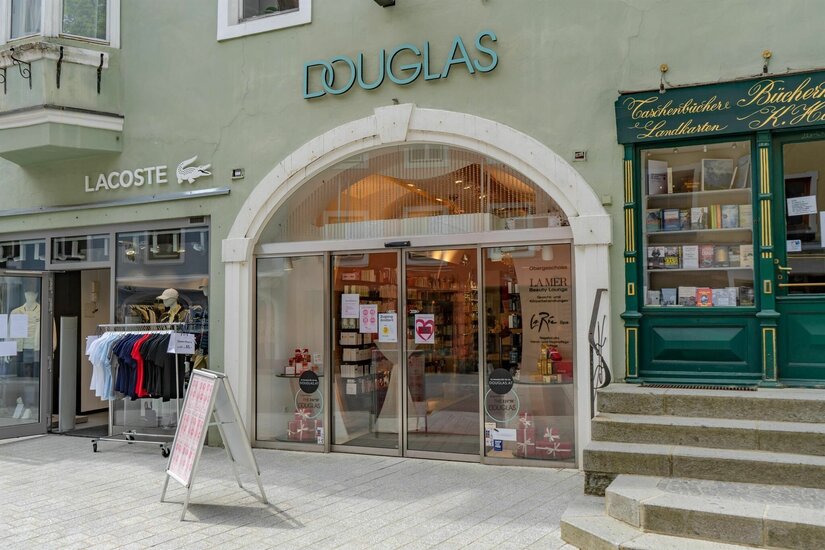 Eingangsbereich einer Douglas-Filiale in einer Fußgängerzone neben einem Lacoste-Shop und einer Buchhandlung.