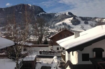 Verschneite Wohnhäuser in einem Alpendorf vor bewaldeten, schneebedeckten Bergen unter blauem Himmel.