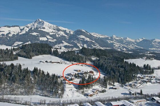 Luftaufnahme einer verschneiten Berglandschaft mit Häusergruppe und Wald vor alpinem Panorama.