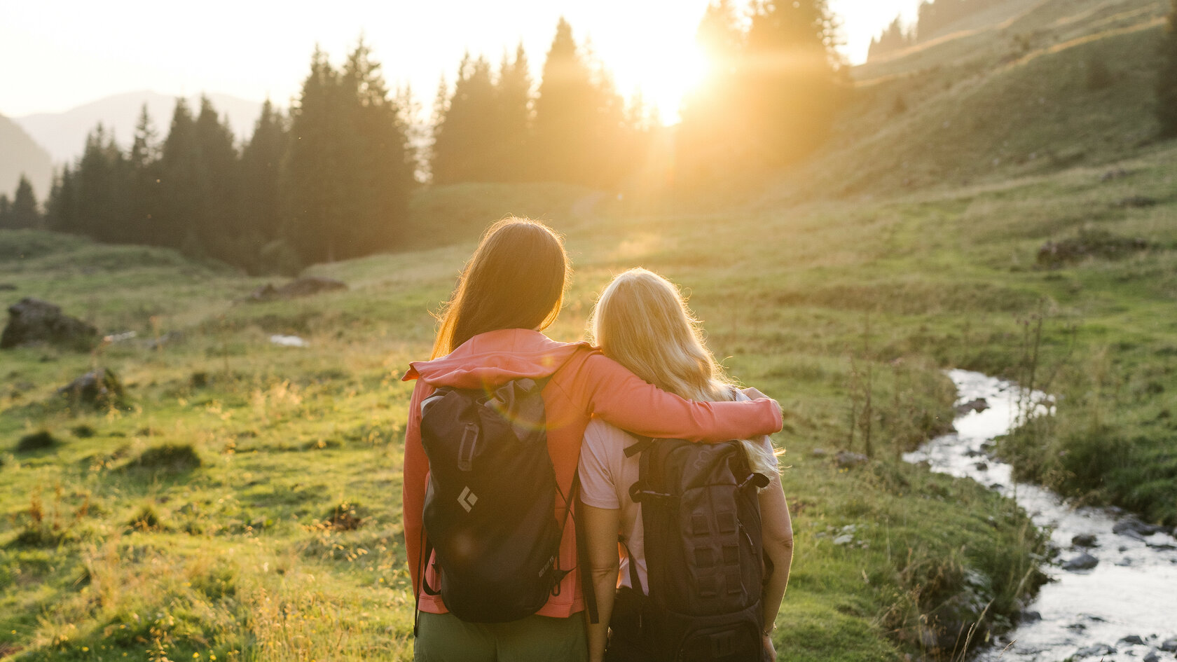 Zwei Wanderer stehen eng umarmt auf einer Wiese, während die Sonne hinter den Bergen untergeht und die Landschaft erleuchtet.