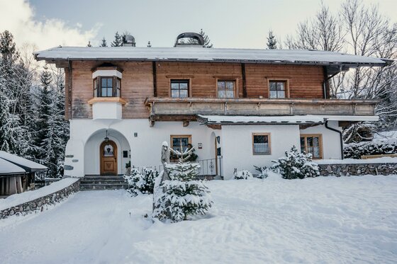 Haus Gantschnigg Außenansicht Winter Zweistöckiges Alpenhaus mit Holzfassade und weißem Erdgeschoss in einer verschneiten Winterlandschaft.