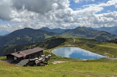 Hornköpflsee mit Berghütte Berghütte neben einem runden Speichersee in einer alpinen Graslandschaft mit Bergpanorama im Hintergrund.