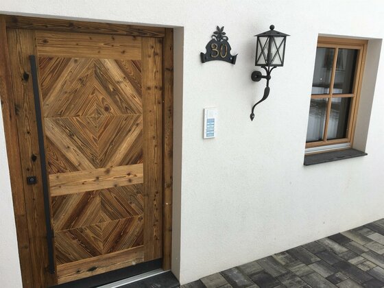 Eingangsbereich eines Hauses mit gemusterter Holztür, Hausnummer 30, Außenleuchte und Fenster.