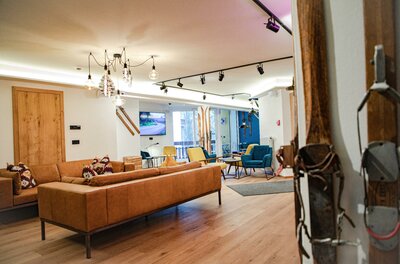 Moderner Loungebereich mit braunem Ledersofa, bunten Sesseln, Holzboden und dekorativen Skiern an der Wand.