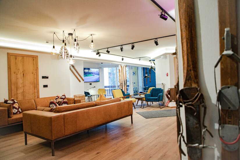 Lounge im Safestay Kitzbühel Moderner Loungebereich mit braunem Ledersofa, bunten Sesseln, Holzboden und dekorativen Skiern an der Wand.