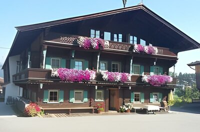 Hölzlhof-Vermietung-Bauernhof-Kitzbühel-Haus Ein traditionelles altes Haus mit mehreren Stockwerken und vielen bunten Blumen auf den Balkonen. Der Himmel ist klar und die Umgebung ist grün.