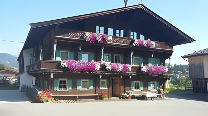 Ein traditionelles altes Haus mit mehreren Stockwerken und vielen bunten Blumen auf den Balkonen. Der Himmel ist klar und die Umgebung ist grün.
