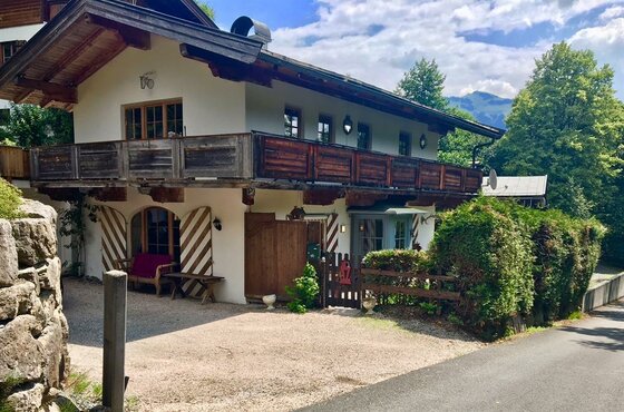 Zweistöckiges alpines Gebäude mit Holzbalkon, weißer Fassade und Fensterläden vor Bäumen.