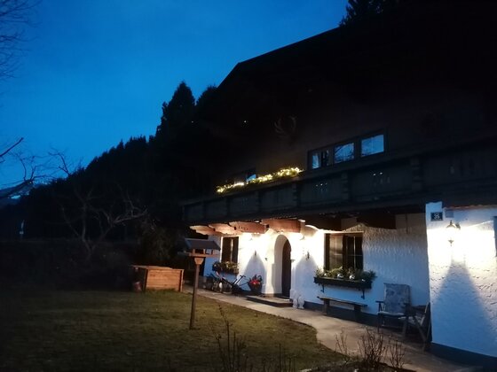 Beleuchtetes alpines Wohnhaus in der Dämmerung mit Balkon und Gartenbereich.