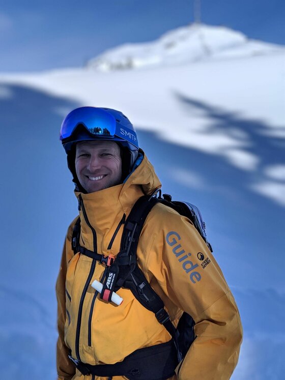 Ein Bergführer in gelber Jacke mit Helm und Lawinenrucksack vor einer verschneiten Bergkulisse.