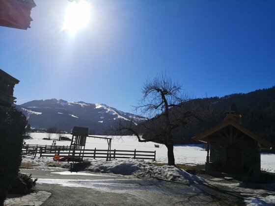 Verschneite Berglandschaft mit kleiner Kapelle, Baum und Spielplatz unter blauem Himmel bei Sonnenschein.