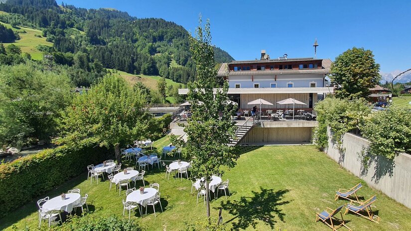 Außenbereich des Gasthof Auwirt mit gedeckten Tischen auf einer Wiese vor dem Gebäude und bewaldeten Bergen.