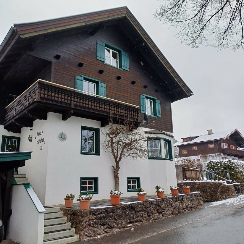 Zweistöckiges alpines Wohnhaus mit dunkler Holzfassade im Obergeschoss und weißem Putz im Erdgeschoss.