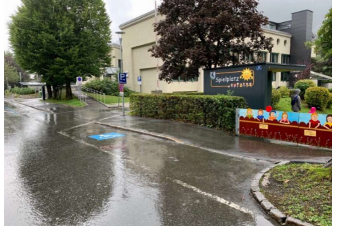 Ein Spielplatz mit einem Schild und bunten Figuren ist sichtbar. Die Straße ist nass vom Regen, und es gibt Bäume in der Umgebung.