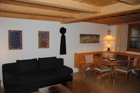 Wohnzimmer mit schwarzem Sofa, Holzeckbank und rundem Esstisch unter einer Balkendecke.