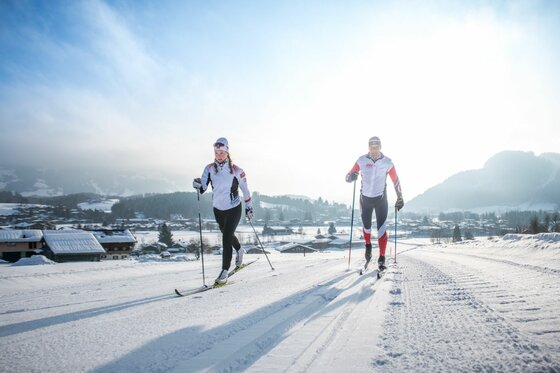 Skilanglauf auf der Lisa Hauser Loipe Zwei Personen beim Skilanglauf auf einer präparierten Loipe vor einer Bergkulisse bei Sonnenschein.