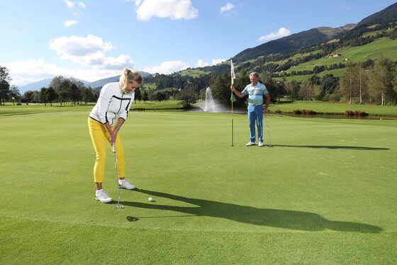 Zwei Personen beim Golfspielen auf einem Green vor einer Bergkulisse mit Wasserfontäne.