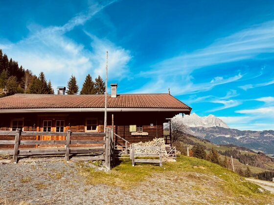 Hütte am Hollenauer Kreuz Rustikale Holzhütte mit Zaun vor einer Bergkulisse unter blauem Himmel.
