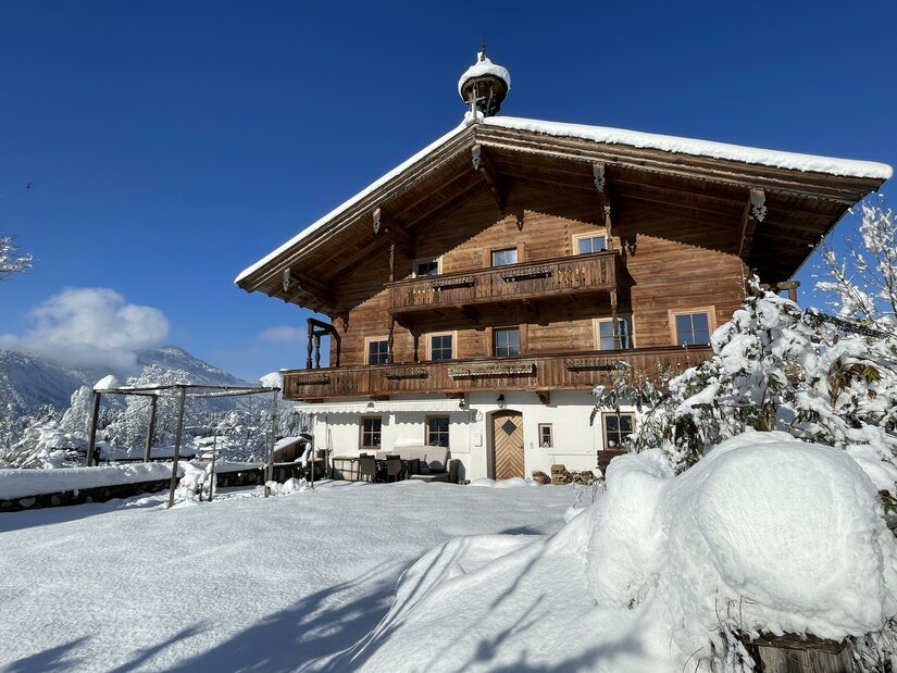 Vorderansicht eines traditionellen Holzhauses mit Balkonen im tiefen Schnee unter blauem Himmel.