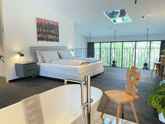 Ein modernes Schlafzimmer mit einem großen Bett und Fußbodenfenstern. Im Vordergrund steht eine Badewanne und ein Holzstuhl.