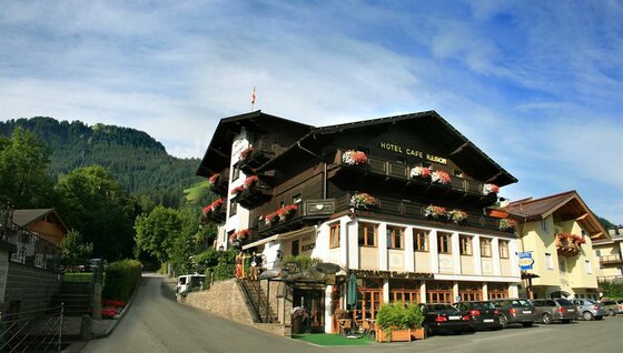 Außenansicht Hotel Cafe Resch Mehrstöckiges Hotelgebäude mit dunkler Holzfassade, Balkonblumen und Parkplatz vor bewaldetem Berg.