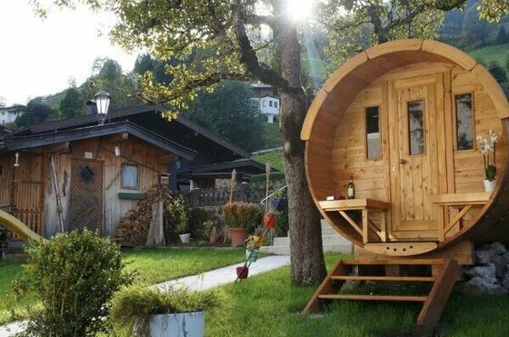 Eine hölzerne Fasssauna im Garten neben einer rustikalen Holzhütte unter Bäumen.